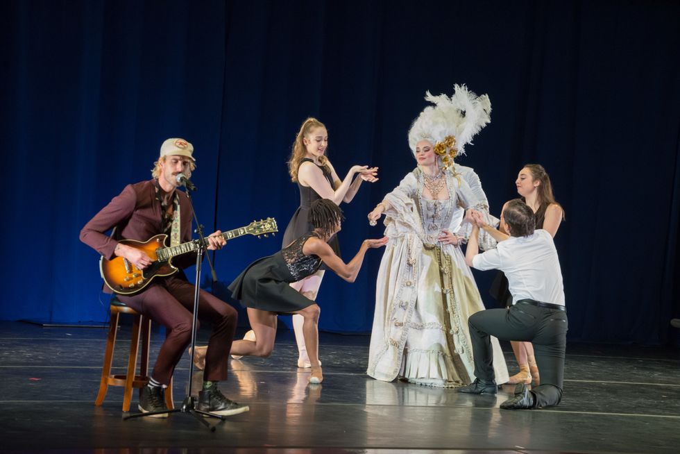 Rayland Baxter sits on a stool while he sings and plays guitar. To his right are three female dancers in black and a male in black and white, surrounding a woman in a tall, white wig and fancy dress.
