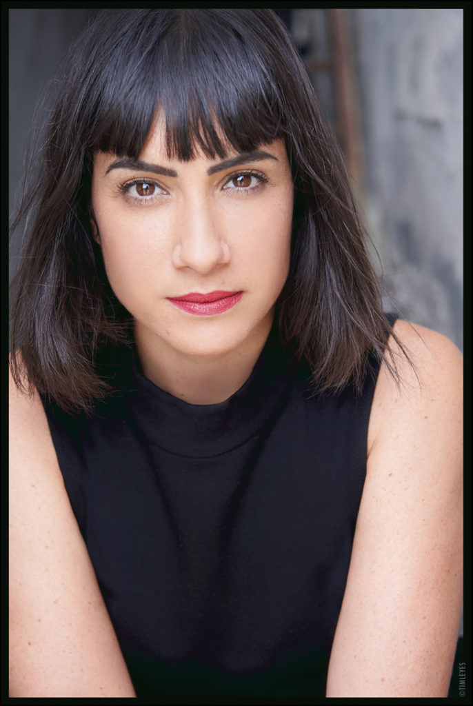 Anisa Tejpar is a young woman with a medium complexion and dark straight hair with cropped bangs. She wears red lipstick and a black sleeveless top and is shown from the waist up, looking into the camera with a neutral expression.